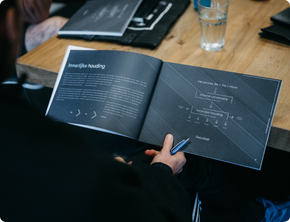 Persoon houdt een open werkboek van The Driven Club boven een tafel